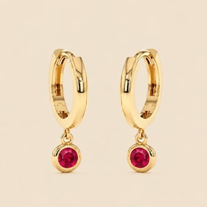 May include: A pair of gold hoop earrings featuring a polished finish and a circular hinge mechanism. Each earring is accented with a single, round, vibrant red gemstone suspended from the base of the hoop, creating a dangling design.