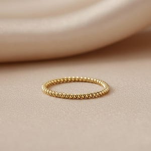 May include: A gold-colored ring with a twisted rope design. The ring is set against a neutral background, highlighting its delicate texture and shine. The ring is a simple band, suitable for everyday wear.