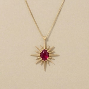 Könnte beinhalten: Goldfarbene Halskette mit einem sternförmigen Anhänger. Der Anhänger hat einen ovalen, roten Edelstein in der Mitte, umgeben von spitzen Strahlen. Die Kette ist fein und goldfarben. Ein auffälliges Schmuckstück.
