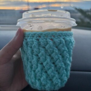 May include: A clear plastic coffee cup with a lid is held in a hand. The cup is wrapped in a light blue crocheted cozy. The background shows a blurred view of a sunset.