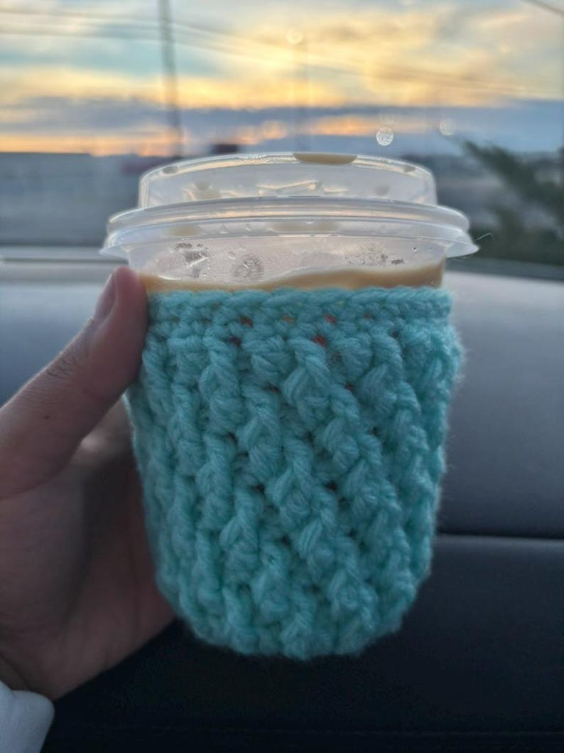 May include: A clear plastic coffee cup with a lid is held in a hand. The cup is wrapped in a light blue crocheted cozy. The background shows a blurred view of a sunset.