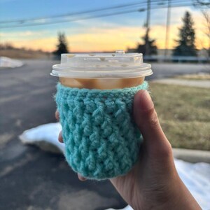 May include: A cup of coffee with a clear plastic lid is held in a hand. The cup is wrapped in a turquoise crocheted cozy. The background shows a sunset with trees and power lines.