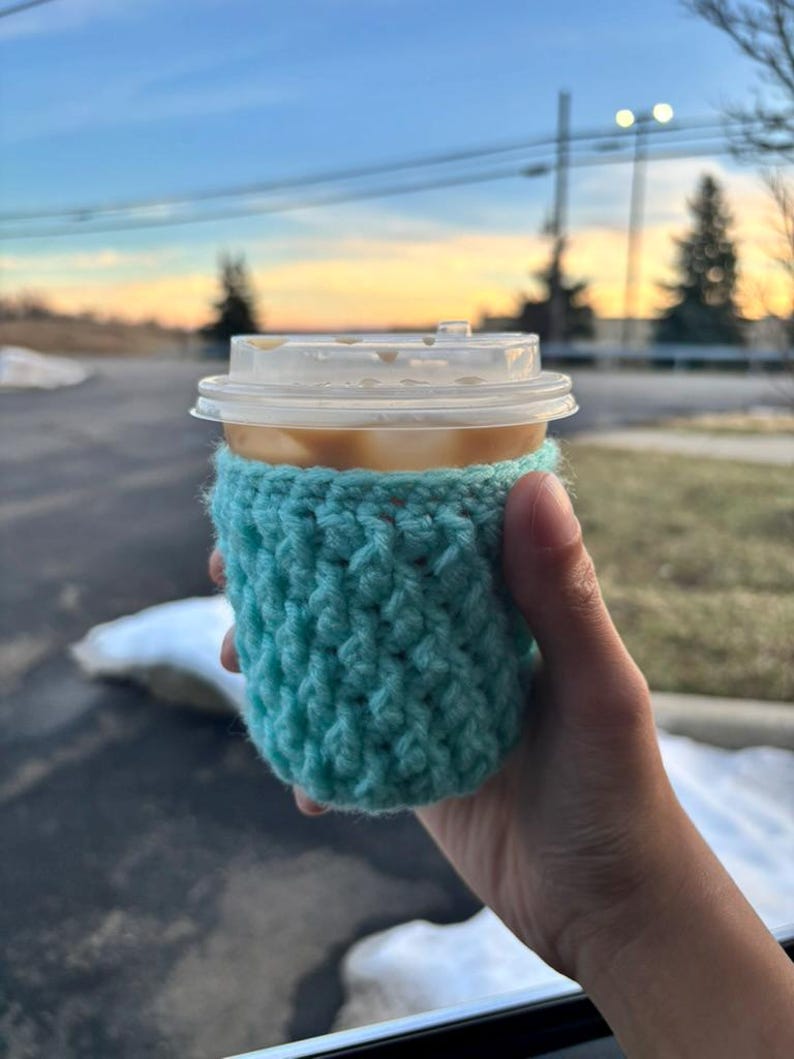 May include: A cup of coffee with a clear plastic lid is held in a hand. The cup is wrapped in a turquoise crocheted cozy. The background shows a sunset with trees and power lines.