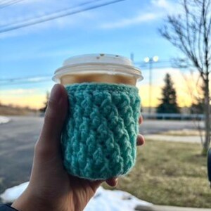 May include: A teal crochet coffee cup sleeve wrapped around a disposable coffee cup with a white lid. The cup is held in a hand, with a blurred outdoor background of a blue sky and trees.