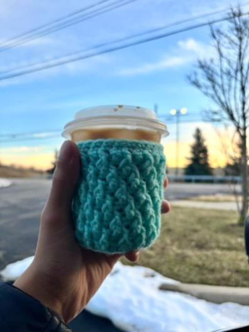 May include: A teal crochet coffee cup sleeve wrapped around a disposable coffee cup with a white lid. The cup is held in a hand, with a blurred outdoor background of a blue sky and trees.
