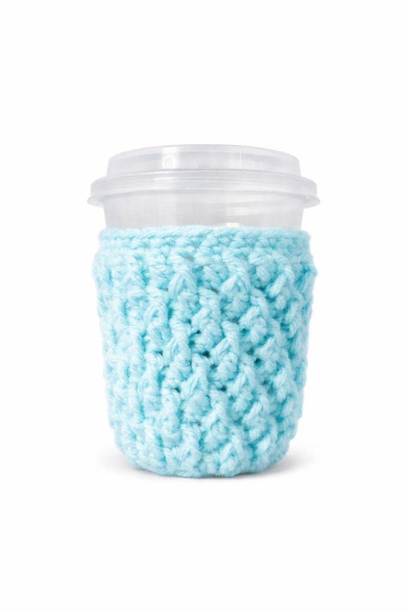 May include: A clear plastic cup with a lid is wrapped in a light blue crocheted cozy. The cozy is made of a textured yarn and fits snugly around the cup, providing insulation and a decorative touch.