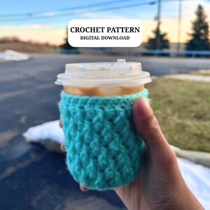 May include: A turquoise crocheted coffee cup cozy, wrapped around a disposable coffee cup with a clear lid. The text "CROCHET PATTERN DIGITAL DOWNLOAD" is in a white rectangle in the background. The cozy is held by a hand.