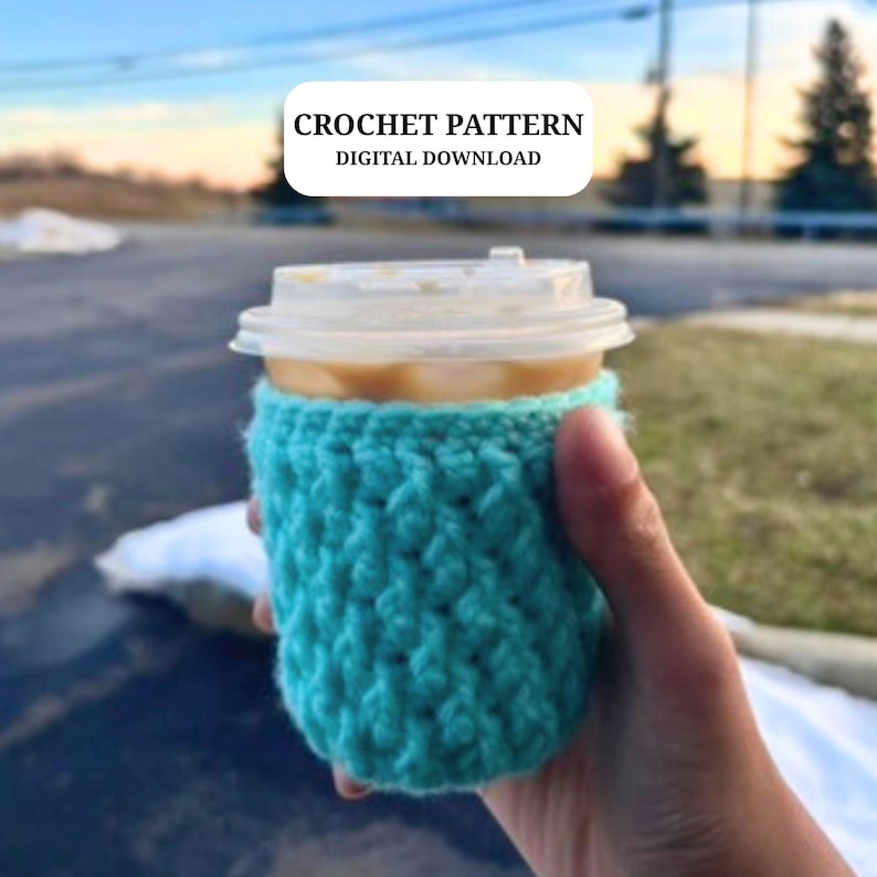 May include: A turquoise crocheted coffee cup cozy, wrapped around a disposable coffee cup with a clear lid. The text "CROCHET PATTERN DIGITAL DOWNLOAD" is in a white rectangle in the background. The cozy is held by a hand.