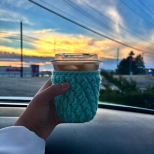 May include: A clear plastic cup filled with a beverage, held in a hand. The cup is wrapped in a teal crocheted cozy. The background shows a colorful sunset with shades of blue, orange, and yellow.