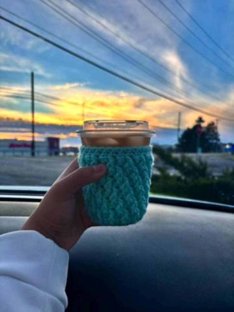 May include: A clear plastic cup filled with a beverage, held in a hand. The cup is wrapped in a teal crocheted cozy. The background shows a colorful sunset with shades of blue, orange, and yellow.