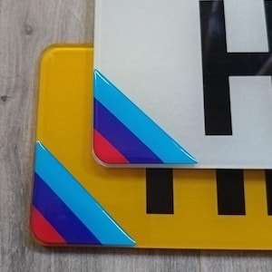 May include: Two vehicle number plates, one yellow and one white, with black characters. Each plate has a triangular corner design with red, blue, and light blue stripes. The plates are set against a light grey wooden background.