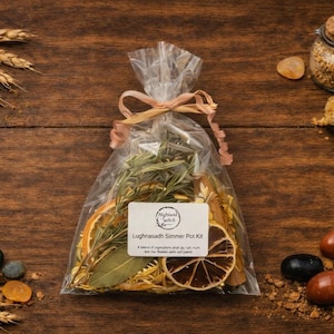 May include: A clear plastic bag containing a Lughnasadh Simmer Pot Kit, featuring dried orange slices, rosemary sprigs, and bay leaves, tied with a light pink ribbon. The white label displays the text Highland Witch and Lughnasadh Simmer Pot Kit.