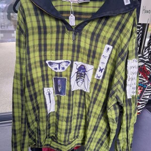 May include: A green and black checked long-sleeved shirt with a black collar and zip. It has insect patches, including butterflies and beetles. The sleeves have black cuffs.