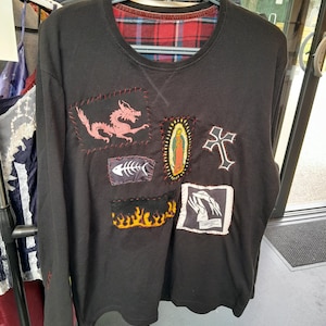 May include: Black long-sleeve shirt with a red and black plaid lining at the neckline. The shirt features several patches, including a pink dragon, a fish skeleton, a religious icon, a cross, flames, and hands.