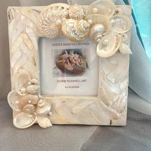 May include: Square photo frame decorated with seashells and faux pearls. The frame is white with a mother-of-pearl finish and features the text "SUZIES SEASHELL WORLD" and "DIVINE SEASHELL ART". The frame is designed for a 4 x 4 inch photo.