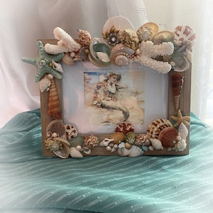 May include: A wooden picture frame decorated with seashells, starfish, and coral. The frame is 5x7 inches and has a mermaid illustration inside. The frame is on a teal fabric background.