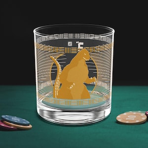 以下が含まれることがあります： 円の中に金のモンスターのイラストが描かれた、透明なロックグラス。縁には金と白の幾何学模様が施されています。グラスは緑色の表面に置かれ、手前にはポーカーチップがあります。