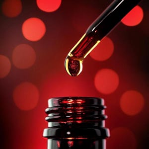 May include: Close-up of a dropper dispensing a golden liquid into a dark glass bottle. The dropper is angled downward, with a single drop suspended at the tip. The background is a blurred red with circular highlights.