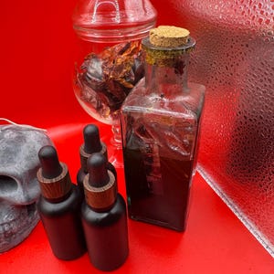 May include: A collection of apothecary-style bottles and jars on a red surface. The image features a clear glass bottle with a cork stopper, several black dropper bottles with wooden tops, and a glass jar filled with dried peppers. A skull is visible on the left.
