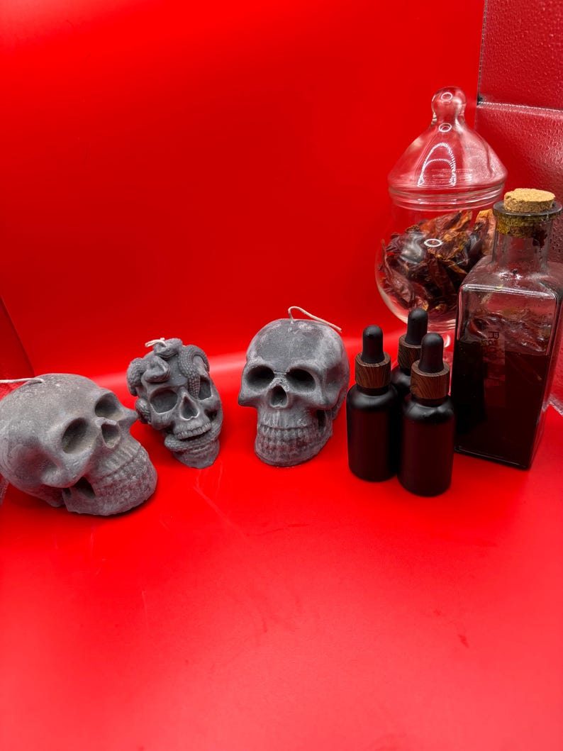May include: A collection of gothic-themed items on a red background. The items include three gray skull candles, several small black bottles with droppers, a glass jar with a lid, and a square glass bottle with a cork.