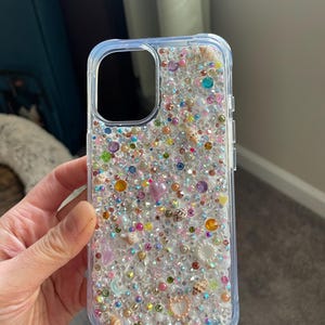 May include: A clear phone case with a glittery design. The case is covered in small, colorful beads, seashells, and pearls.