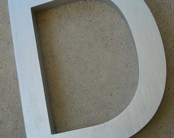 May include: A silver metal letter D with a brushed finish. The letter is standing on a concrete surface.