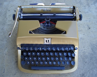 Op de afbeelding: Een vintage goudkleurige typemachine met zwarte toetsen en het woord "de luxe" bovenaan. De toetsen zijn in een QWERTY-indeling geplaatst. De typemachine staat op een grijze ondergrond.