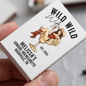 May include: A white rectangular matchbox featuring a graphic of a person in a cowboy hat and red boots sitting in a large margarita glass. The text reads WILD WILD WIFE, MELISSA'S COWGIRL BACHELORETTE NASHVILLE, TN, and EST. 20XX in black lettering.