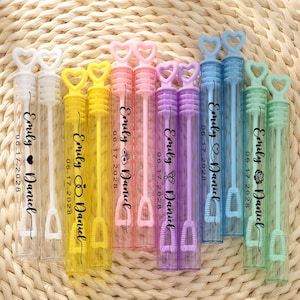 May include: A collection of twelve cylindrical bubble wands in white, yellow, pink, purple, blue, and mint green. Each wand features a heart-shaped handle and is printed with the names Emily and Daniel and the date 06.17.2028 in black text.