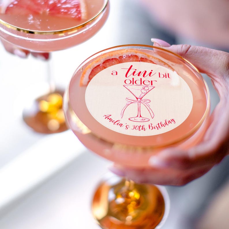 Puede incluir: Un vaso de c&oacute;ctel rosa con un posavasos redondo de papel blanco que dice "a tiny bit older" con una ilustraci&oacute;n de copa de martini y el texto "Amelia's 30th Birthday".