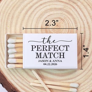 May include: A white matchbox, measuring 2.3 inches long and 1.4 inches wide, with the text "the PERFECT MATCH" and the names Jason & Anna, and the date 04.22.2026. The matches have white tips and are displayed on a wooden surface.