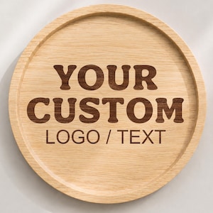 K&ouml;nnte beinhalten: Ein rundes, helles Holztablett mit erh&ouml;htem Rand. In der Mitte des Tabletts befindet sich eine dunkelbraune Gravur mit dem Text "YOUR CUSTOM LOGO / TEXT" in einer fetten, serifenlosen Schrift, die die nat&uuml;rliche Holzmaserung hervorhebt.