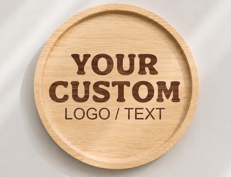 K&ouml;nnte beinhalten: Ein rundes, helles Holztablett mit erh&ouml;htem Rand. In der Mitte des Tabletts befindet sich eine dunkelbraune Gravur mit dem Text "YOUR CUSTOM LOGO / TEXT" in einer fetten, serifenlosen Schrift, die die nat&uuml;rliche Holzmaserung hervorhebt.