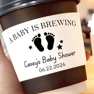 May include: A black paper coffee cup with a white paper sleeve. The sleeve reads "A BABY IS BREWING" above two black footprints, stars, and "Casey's Baby Shower 06.22.2026". Another cup is visible in the background.