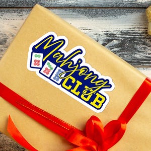 May include: A sticker reading "Mahjong Club" in a blue and yellow design, placed on a brown gift wrapped with a red ribbon. The image includes a gold ornament.