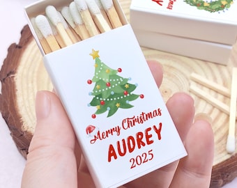 May include: A white matchbox with a Christmas tree design, the text "Merry Christmas AUDREY 2025" in red. The match heads are white. The box is held in a hand.