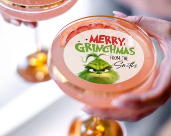 May include: A pink cocktail in a stemmed glass, garnished with a slice of grapefruit. The glass has a sticker with the Grinch and the text "MERRY GRINCHMAS FROM THE Smiths". Another similar cocktail is visible in the background.