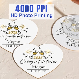 以下が含まれることがあります： シャンパンの乾杯と「2025 Congratulations Megan I DID IT」の文字が描かれた、金色のデザインが施された白いハート型のコースター3枚。
