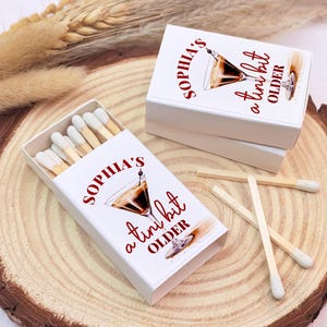 May include: White matchbox with red text that reads "Sophia's a tiny bit older" and a graphic of a cocktail glass. The matchbox is sitting on a wooden surface with a few matches next to it.