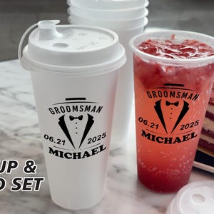 May include: A set of two white cups with lids. The cups have black text that reads "Groomsman 06.21 2025 Michael" and a tuxedo graphic. One cup is filled with a red beverage.