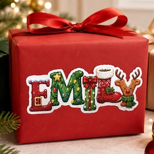 May include: A rectangular gift box wrapped in solid red paper with a large red satin ribbon bow on top. A sticker on the front displays the name EMILY in festive holiday-themed letters, featuring a Santa boot, Christmas tree, and reindeer.