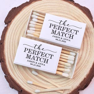 May include: Two white matchboxes with the text "the PERFECT MATCH" and the names Jason & Anna, with the date 04.22.2026. The matchboxes are on a wooden slice, with several matches visible.