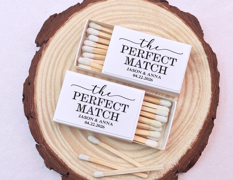 May include: Two white matchboxes with the text "the PERFECT MATCH" and the names Jason & Anna, with the date 04.22.2026. The matchboxes are on a wooden slice, with several matches visible.