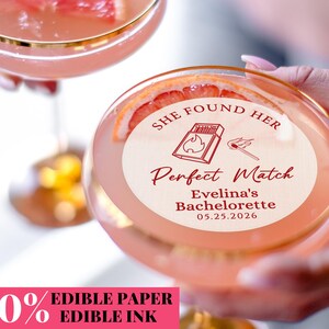 Könnte beinhalten: Zwei Cocktailgläser mit rosa Getränk und Grapefruit-Scheiben-Garnitur. Ein Glas hat einen Aufkleber mit der Aufschrift "She Found Her Perfect Match Evelina's Bachelorette 05.25.2026" mit einer Streichholz- und Streichholzschachtel-Illustration.