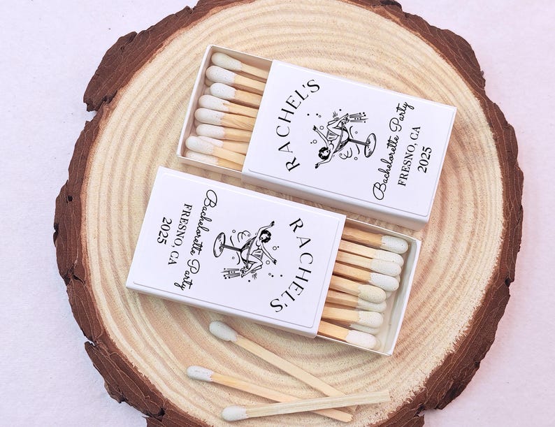 May include: Two white matchboxes with black text and a graphic of a woman holding a martini glass. The text on the boxes reads "Rachel's Bachelorette Party Fresno, CA 2025".