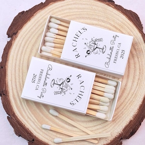 May include: Two white matchboxes with black text and a graphic of a woman holding a martini glass. The text on the boxes reads "Rachel's Bachelorette Party Fresno, CA 2025".