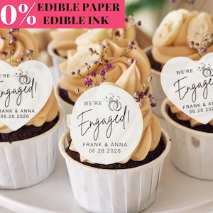 May include: Cupcakes with tan frosting, topped with heart-shaped edible paper that reads "We're Engaged!" with names and a date. The cupcakes are in white paper cups and decorated with small purple flowers. The text "100% Edible Paper Edible Ink" is in the top left.