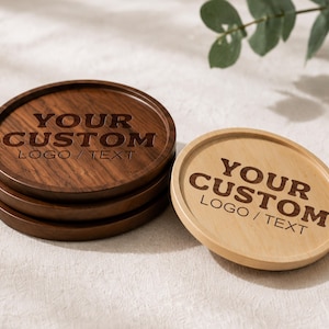 K&ouml;nnte beinhalten: Ein Stapel aus drei dunkelbraunen runden Holzuntersetzern und einem hellbraunen runden Holzuntersetzer. Jeder Untersetzer hat einen erh&ouml;hten Rand und ist mit dem Text YOUR CUSTOM LOGO / TEXT in einer fetten Schrift auf der Fl&auml;che graviert.