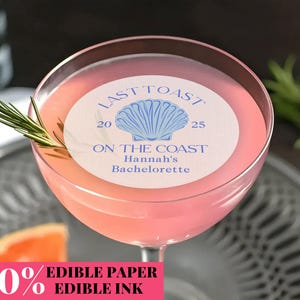 Op de afbeelding: Een roze cocktail in een glas met steel, gegarneerd met rozemarijn. De drank heeft een cirkelvormig eetbaar papier met de tekst "LAST TOAST ON THE COAST Hannah's Bachelorette" en een schelpmotief. Een schijfje grapefruit is zichtbaar.