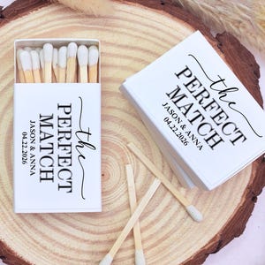 May include: White matchboxes with the text "the PERFECT MATCH" and the names Jason & Anna, along with the date 04.22.2026. The matches have white tips and are displayed on a wooden surface.
